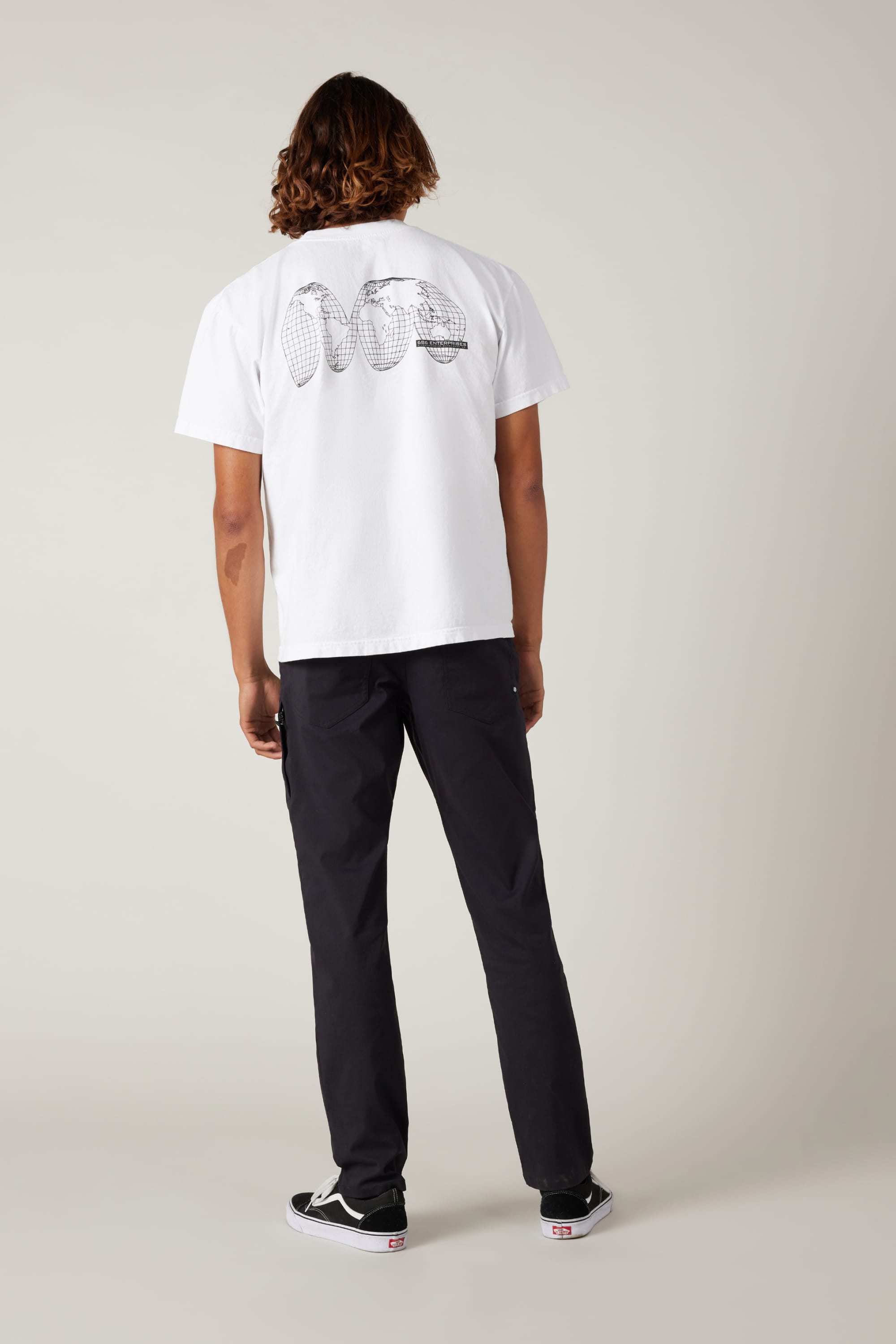A person with shoulder-length hair stands facing away, wearing a white T-shirt with a black graphic on the back, 686 Men's Everywhere® Pant - Slim Fit by 686, and black shoes, against a plain light background.