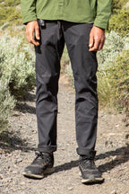 A person stands on a rocky trail, wearing 686 Men's Everywhere® Pant - Slim Fit. Only their lower half is visible among green shrubs, paired with black sneakers and a green jacket.