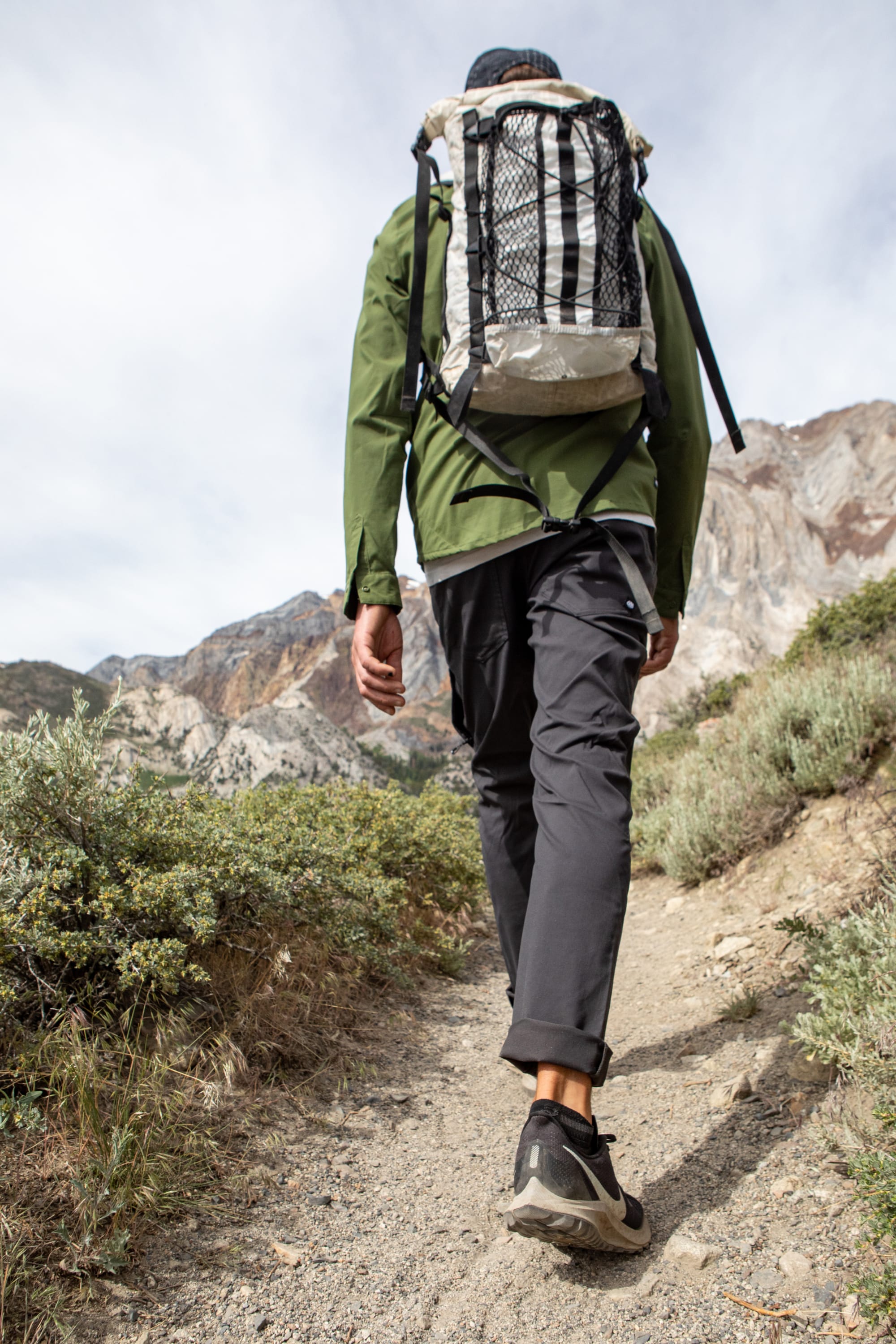 A hiker in a green jacket, backpack, and 686 Men's Everywhere® Pant - Slim Fit treks up a dirt trail lined with shrubs and rocks, mountains ahead under a partly cloudy sky. The scene is viewed from behind and below.