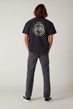 A person with shoulder-length hair faces away, wearing a black 686 graphic tee, 686 Men's Everywhere® Pant - Slim Fit in gray, and black-and-white sneakers, set against a plain light gray background.