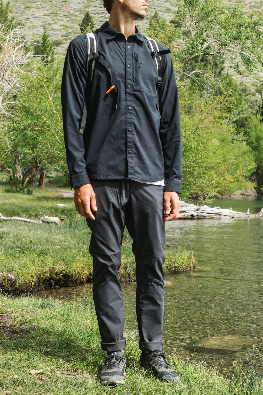 A person in a dark long-sleeve shirt, 686 Men's Everywhere® Pant - Slim Fit, hiking boots, and a backpack stands on grass by a lake with trees and hills behind them. Their face is not fully visible.
