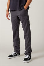 A person wearing 686 Men's Everywhere® Pant - Slim Fit in dark gray, paired with a dark t-shirt and black-and-white sneakers, stands against a plain light background. Only their body from the shoulders down is visible, with hands relaxed at their sides.