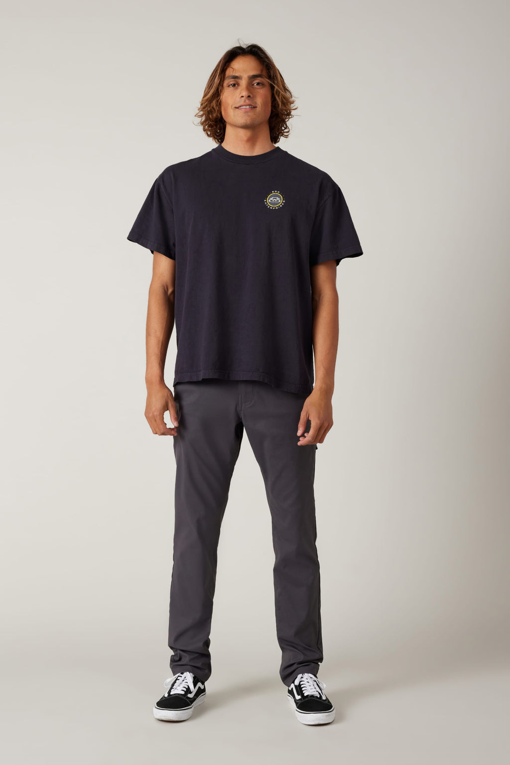 A person faces forward against a light background, wearing a loose black t-shirt, 686 Men's Everywhere® Pant - Slim Fit in gray by 686, and black sneakers with white soles.