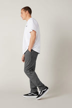 A person stands in profile against a plain light background, wearing a white T-shirt, 686 Men's Everywhere® Pant - Slim Fit from 686 in gray, and black Vans sneakers with white soles. One hand is in their pocket as they look ahead neutrally.