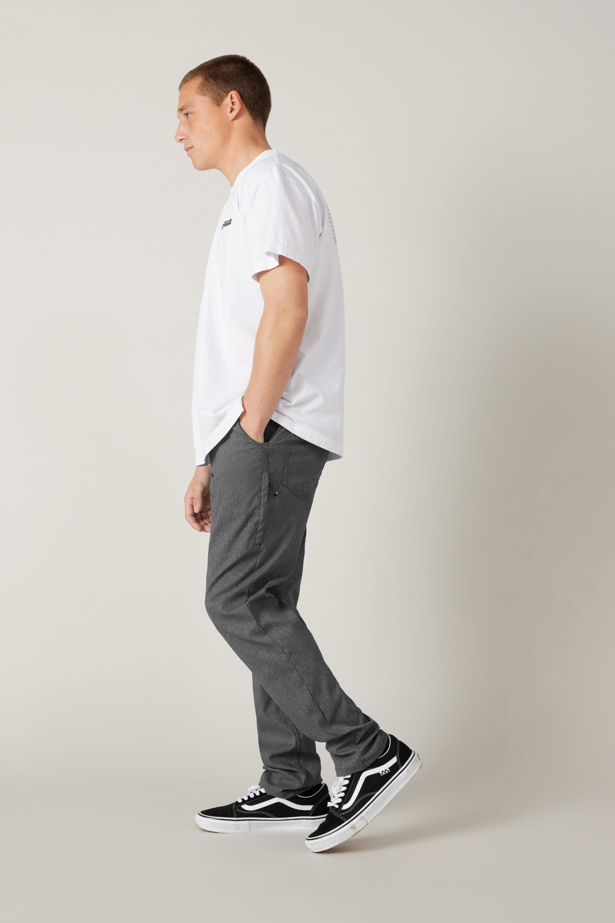 A person stands in profile against a plain light background, wearing a white T-shirt, 686 Men's Everywhere® Pant - Slim Fit from 686 in gray, and black Vans sneakers with white soles. One hand is in their pocket as they look ahead neutrally.