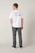 A man stands facing away, wearing a white T-shirt with a black graphic on the back, grey 686 Men's Everywhere® Pant - Slim Fit, and black Vans sneakers, against a plain light-colored background.