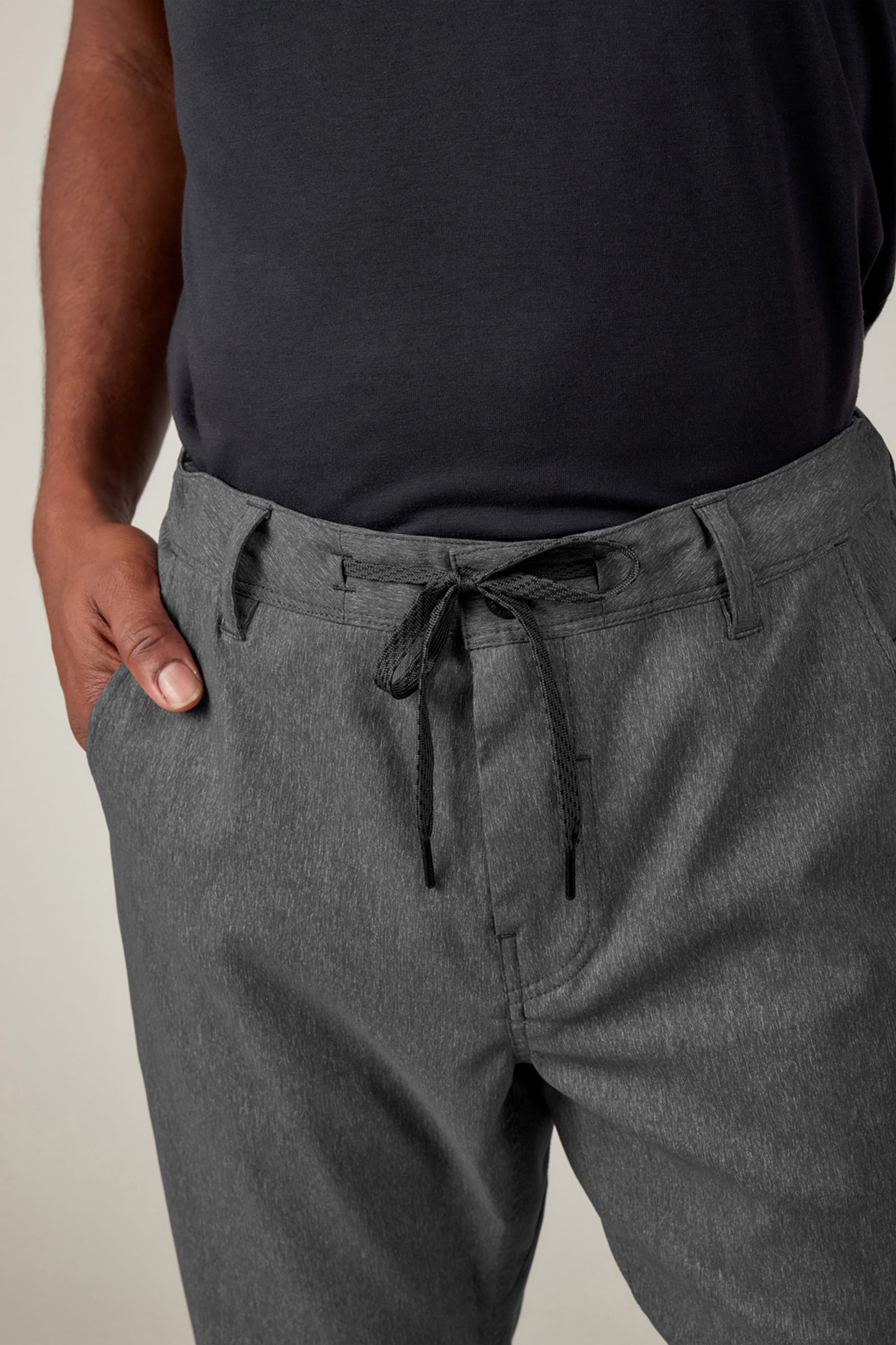 A person wearing a black t-shirt and 686 Men's Everywhere® Pant - Slim Fit in gray, featuring belt loops, a tied waist string, and one hand in their pocket.
