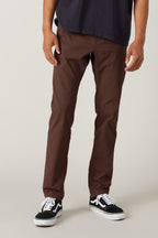 A person in 686 Men's Everywhere® Pant - Slim Fit (brown) by 686, with a dark shirt and black-and-white sneakers, stands against a plain light background. Their hands are at their sides and their face is not visible.