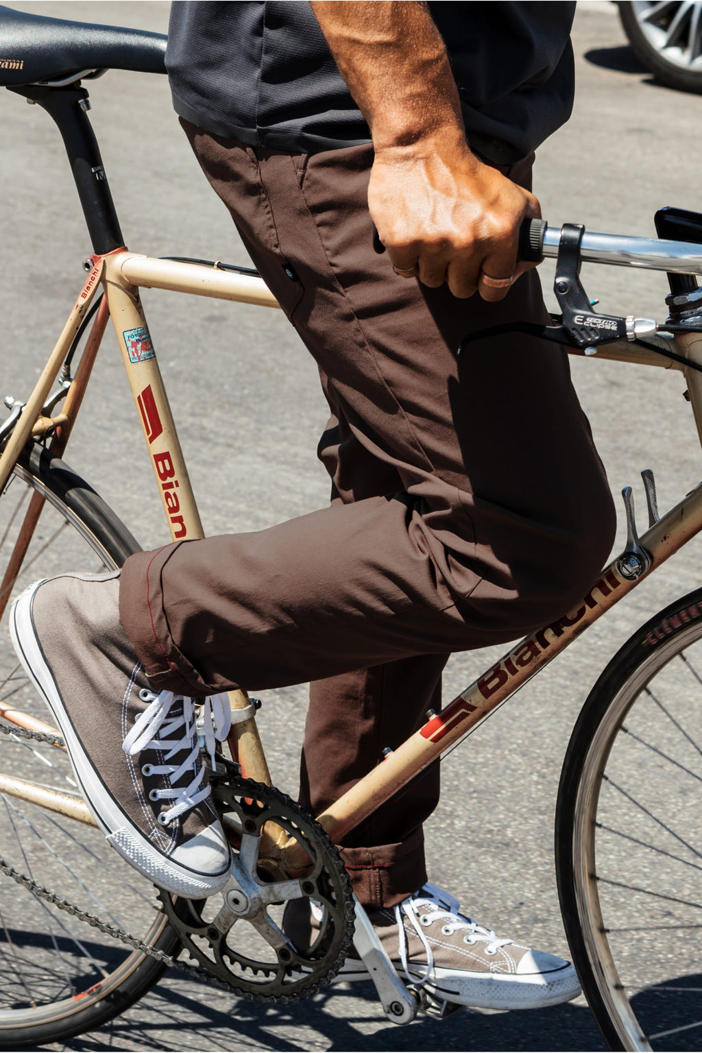 A person in 686 Men's Everywhere® Pant - Slim Fit, stain-resistant brown, and gray sneakers stands beside a beige Bianchi bike, holding the handlebars with one foot on a pedal and the other on the ground.