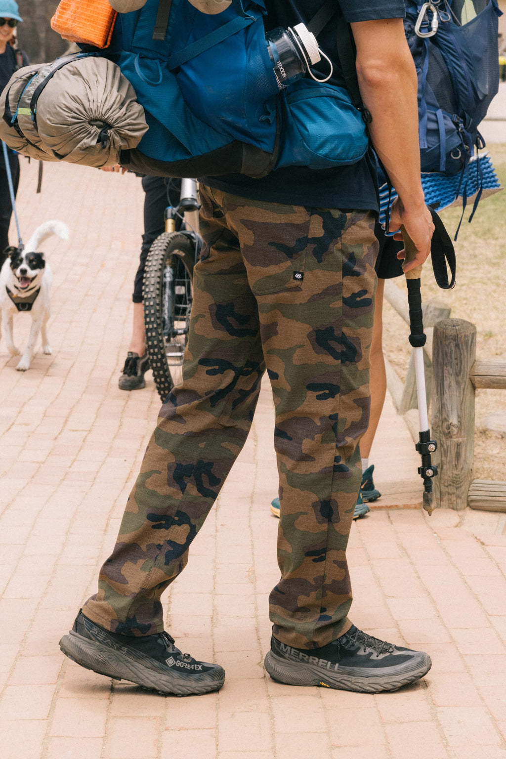 Wearing 686 Men's Everywhere® Pant - Slim Fit, black hiking shoes, and a large blue backpack, a person stands on a paved path with hiking gear. A dog and another person with a bicycle appear in the background.