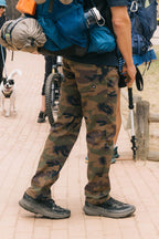 Wearing 686 Men's Everywhere® Pant - Slim Fit, black hiking shoes, and a large blue backpack, a person stands on a paved path with hiking gear. A dog and another person with a bicycle appear in the background.