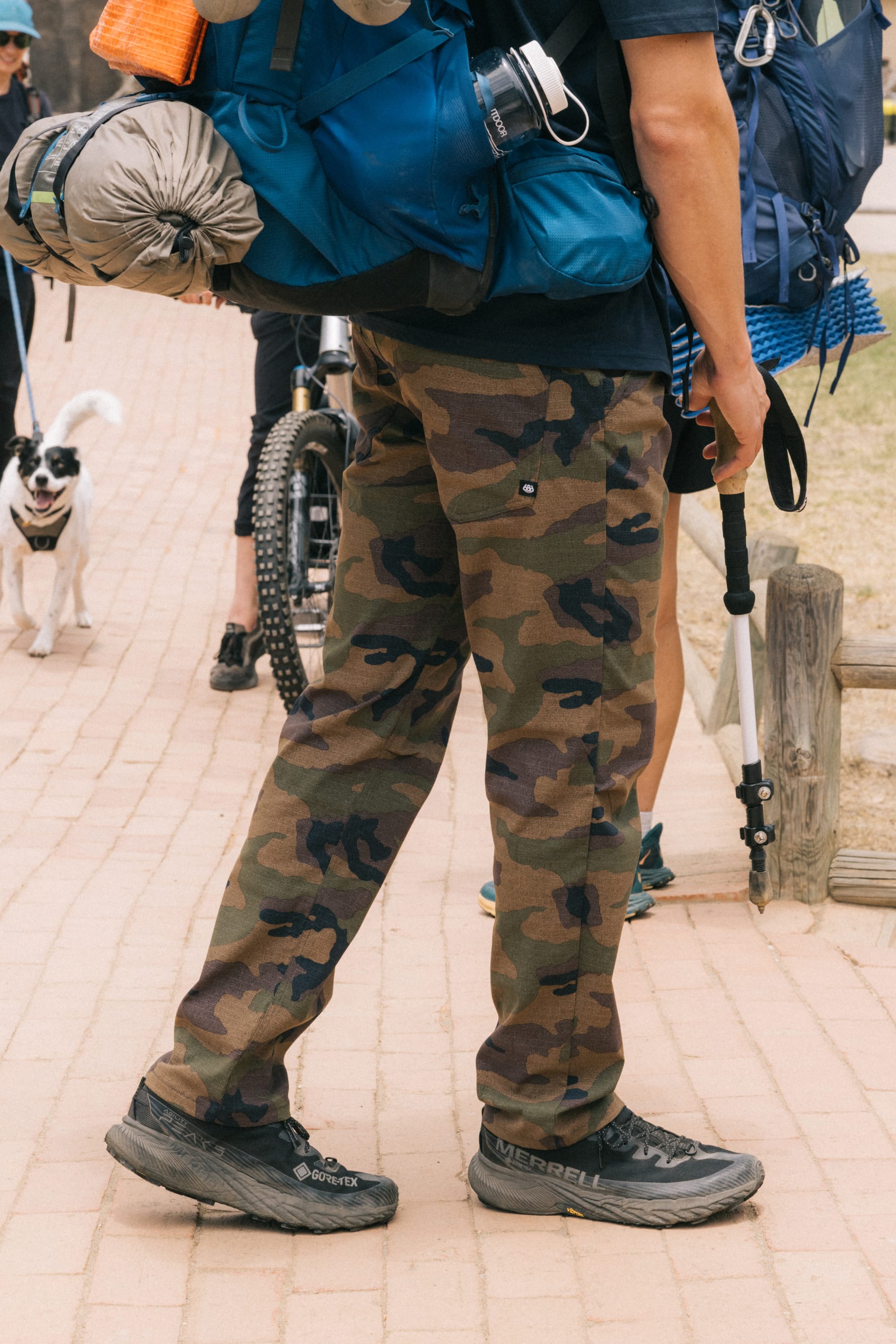 Wearing 686 Men's Everywhere® Pant - Slim Fit, black hiking shoes, and a large blue backpack, a person stands on a paved path with hiking gear. A dog and another person with a bicycle appear in the background.