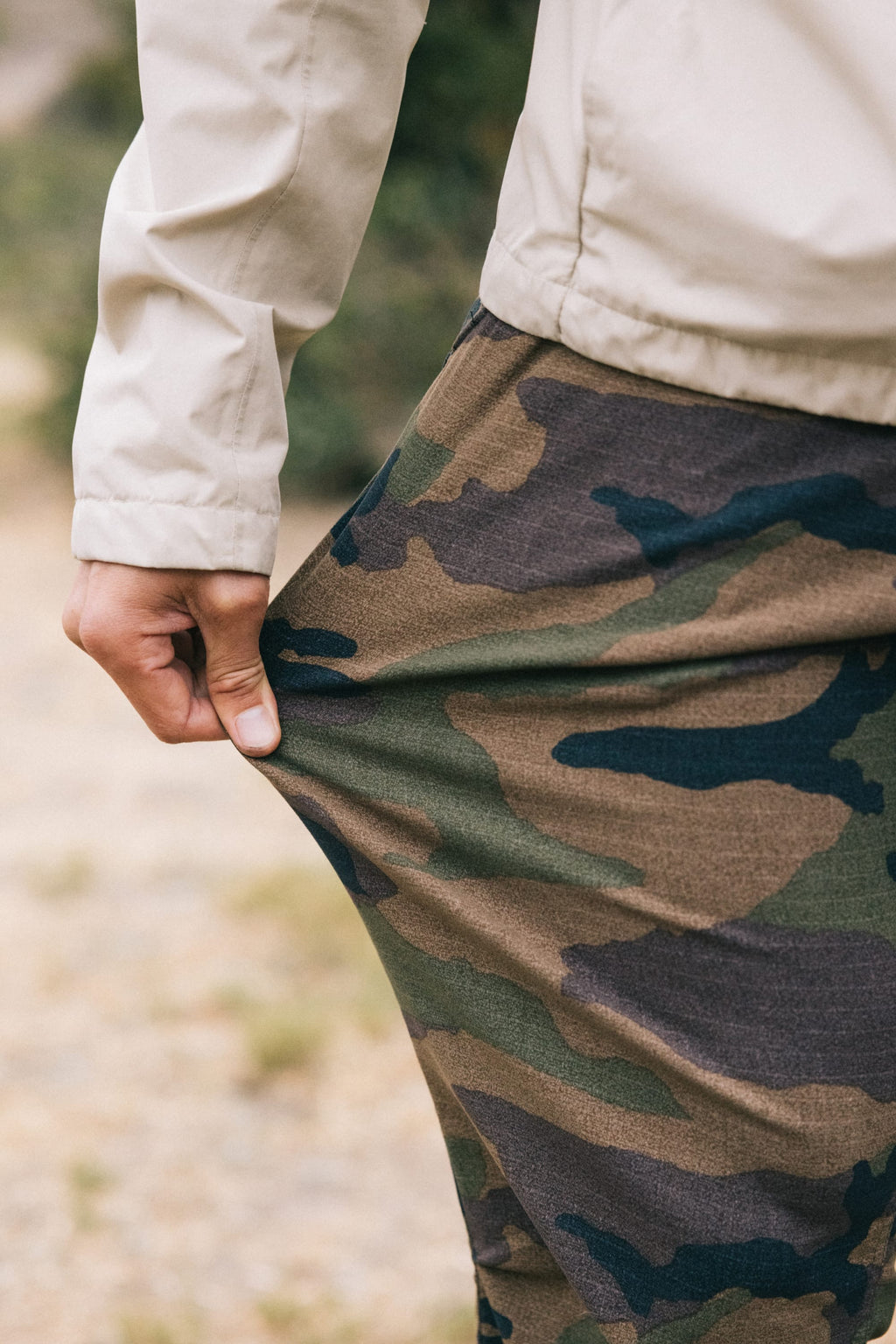Outdoors, a person stretches the fabric of their 686 Men's Everywhere® Pant - Slim Fit in camouflage, showcasing the breathable, stain-resistant material's texture and flexibility.