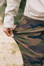 Outdoors, a person stretches the fabric of their 686 Men's Everywhere® Pant - Slim Fit in camouflage, showcasing the breathable, stain-resistant material's texture and flexibility.