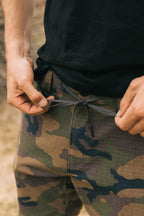 A person, visible from the waist down, ties the drawstring on breathable, stain-resistant 686 Men's Everywhere® Pant - Slim Fit camouflage pants while wearing a black shirt.