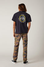 A person with shoulder-length hair stands facing away, wearing a black t-shirt with a circular logo and GBG ENTERPRISE text, 686 Men's Everywhere® Pant - Slim Fit by 686 in camouflage, and black Vans sneakers, against a neutral background.