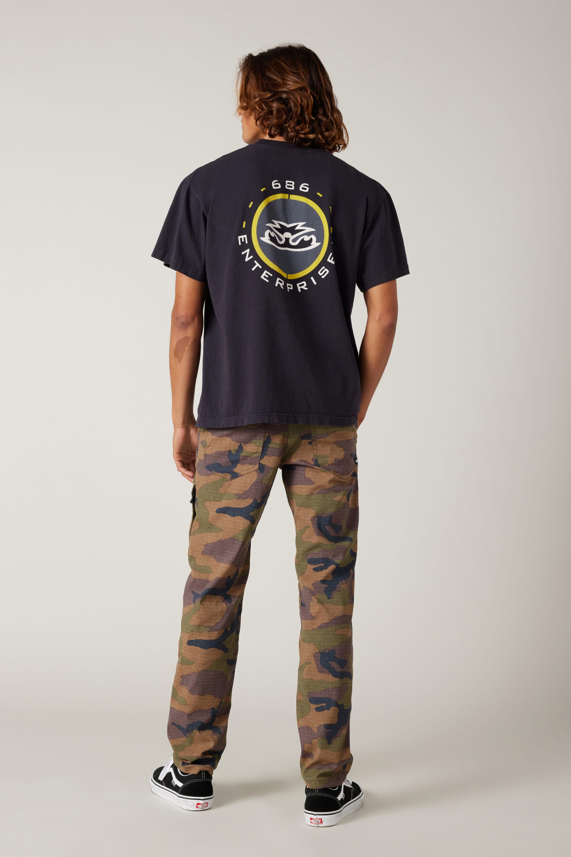 A person with shoulder-length hair stands facing away, wearing a black t-shirt with a circular logo and GBG ENTERPRISE text, 686 Men's Everywhere® Pant - Slim Fit by 686 in camouflage, and black Vans sneakers, against a neutral background.