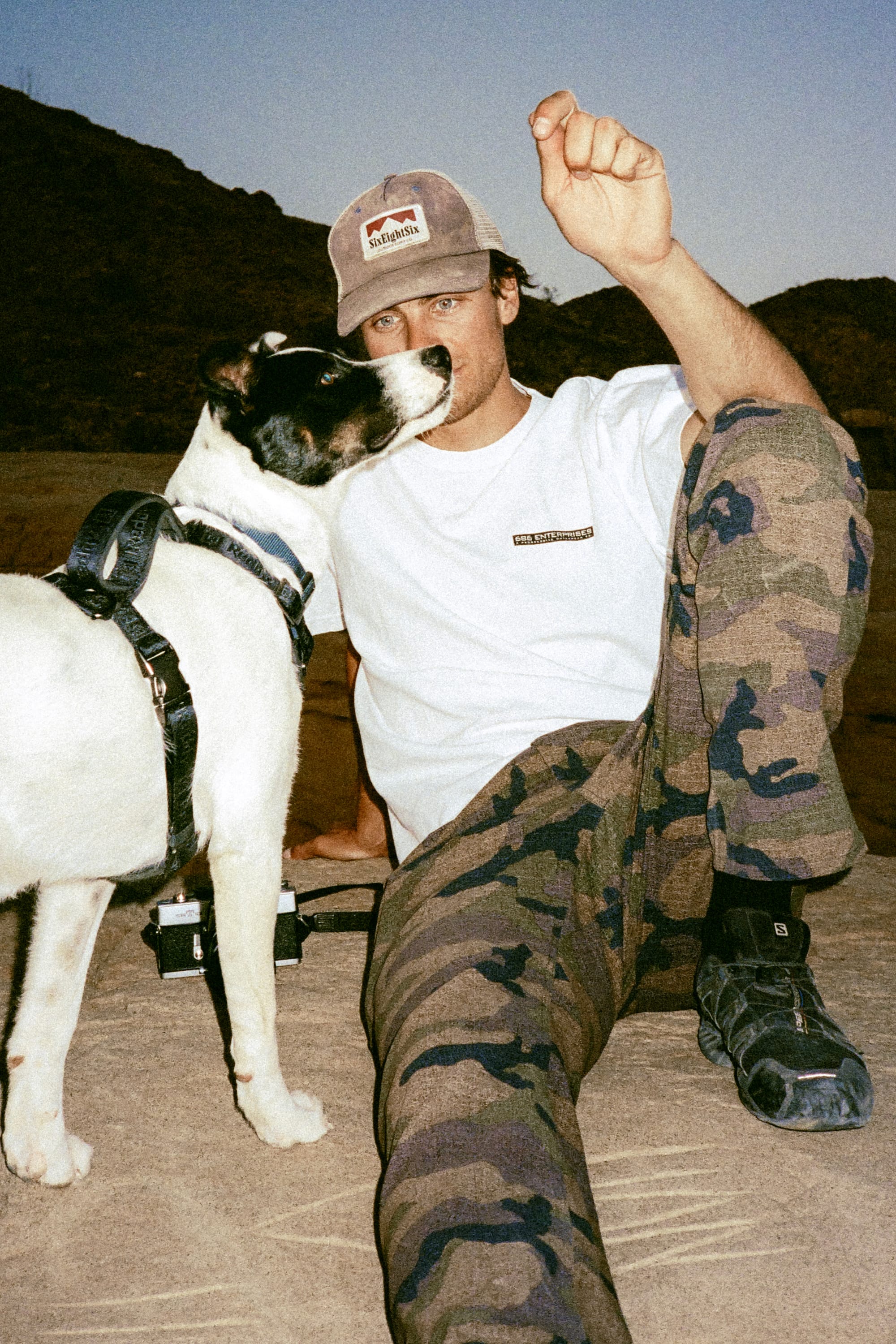 A man in 686 Men's Everywhere® Pant - Slim Fit and a cap sits outdoors, holding something up. A black and white dog in a harness stands beside him, sniffing his face, with hills visible in the background.