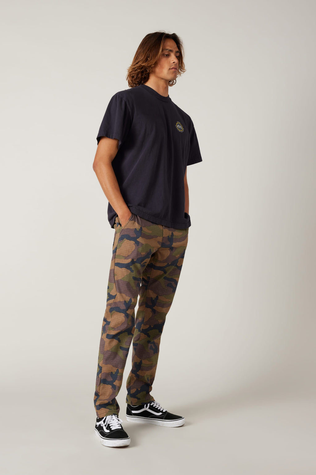 A person with long hair wears a loose black T-shirt, 686 Men's Everywhere® Pant - Slim Fit, and black Vans sneakers. They stand against a plain, light background with hands in their pockets, looking slightly to the side.