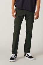 A person wearing 686 Men's Everywhere® Pant - Slim Fit in dark green, a dark shirt, and black-and-white sneakers stands against a plain light background. The photo is cropped from the shoulders down.