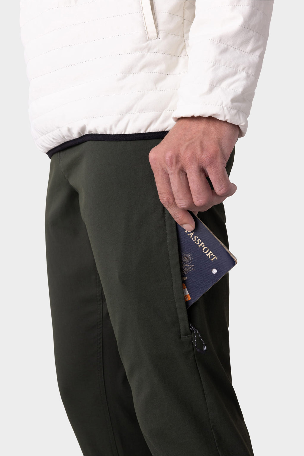 Wearing 686 Men's Everywhere® Pant - Slim Fit in dark green and a white jacket, a person places a navy blue passport into the pant's zippered side pocket. Only their lower torso and hand are visible.