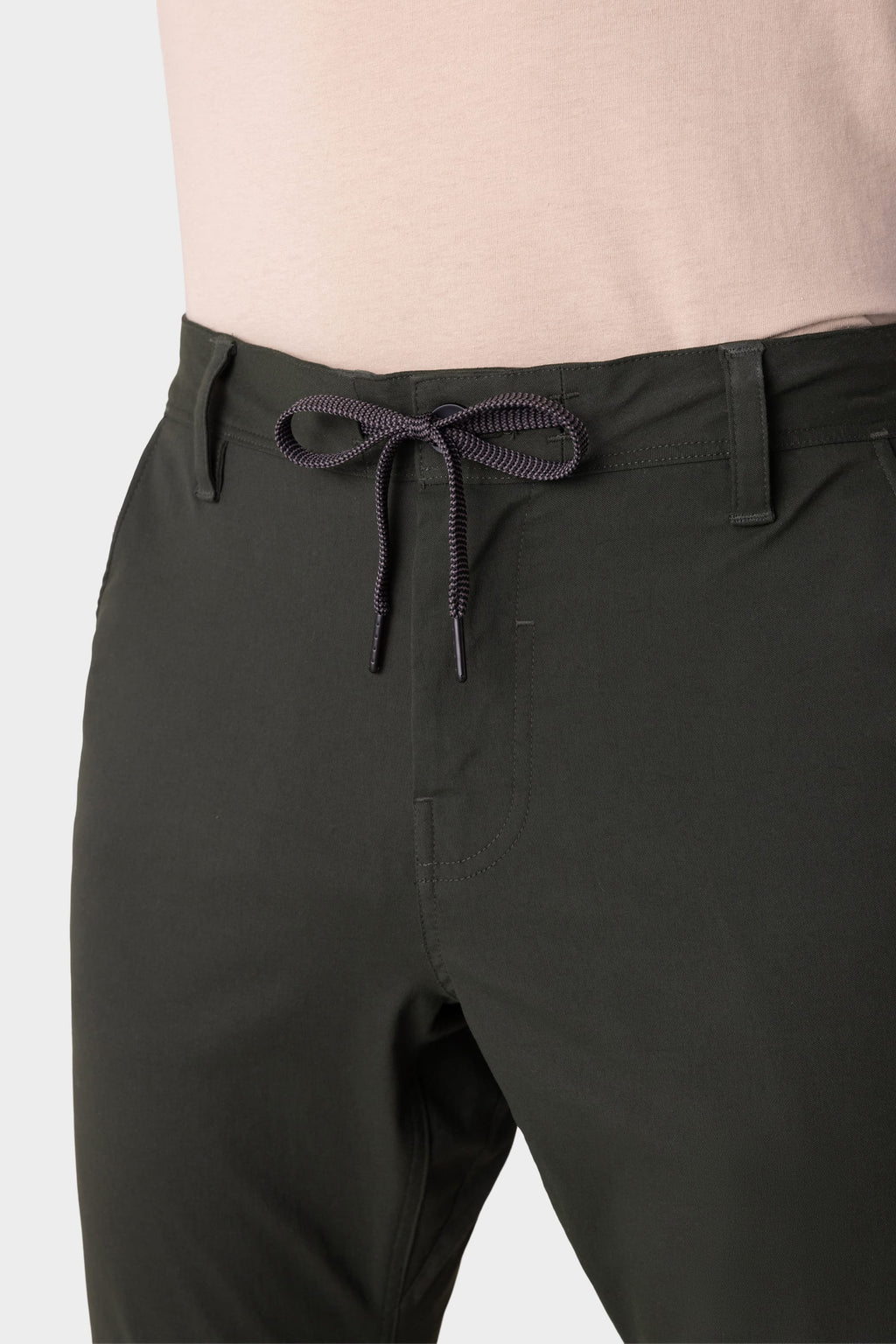 A man wears 686 Men's Everywhere® Pant - Slim Fit in dark green, featuring a front bow-tied drawstring, paired with a light pink shirt, shown from the waist down.