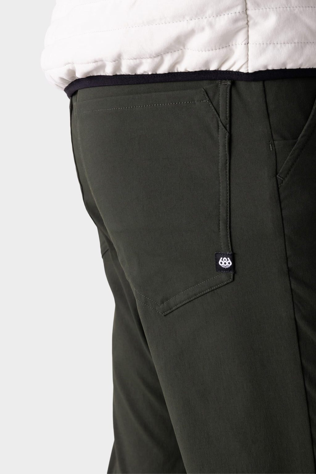 Close-up of a person wearing the 686 Men's Everywhere® Pant - Slim Fit in dark green, paired with a white jacket. The image highlights the breathable pants’ back pocket and the small black 686 tag with a white logo.