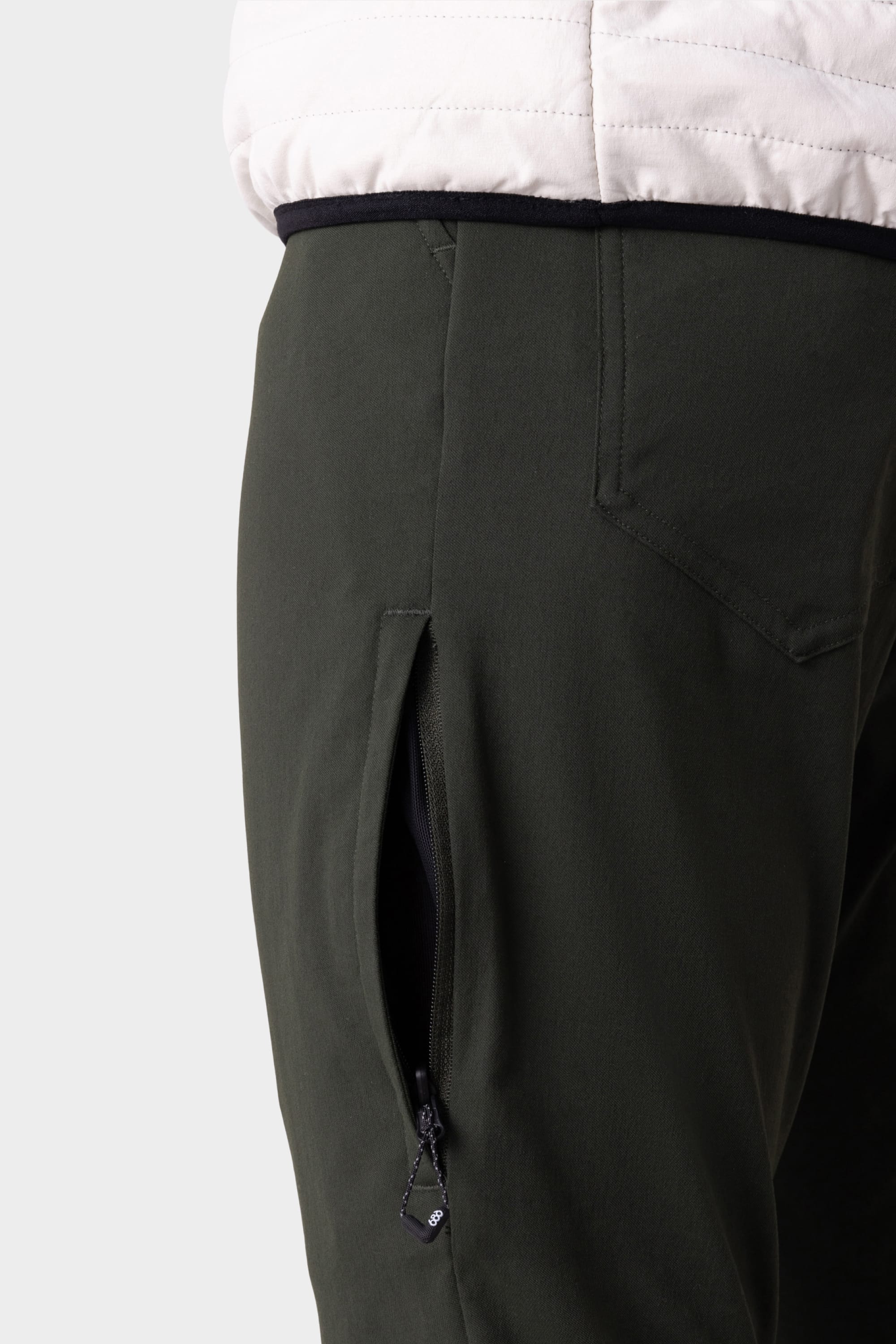 Close-up of a person wearing 686 Men's Everywhere® Pant - Slim Fit in dark green, featuring a visible zippered side pocket. The image highlights the hip and upper leg, paired with a white quilted jacket.