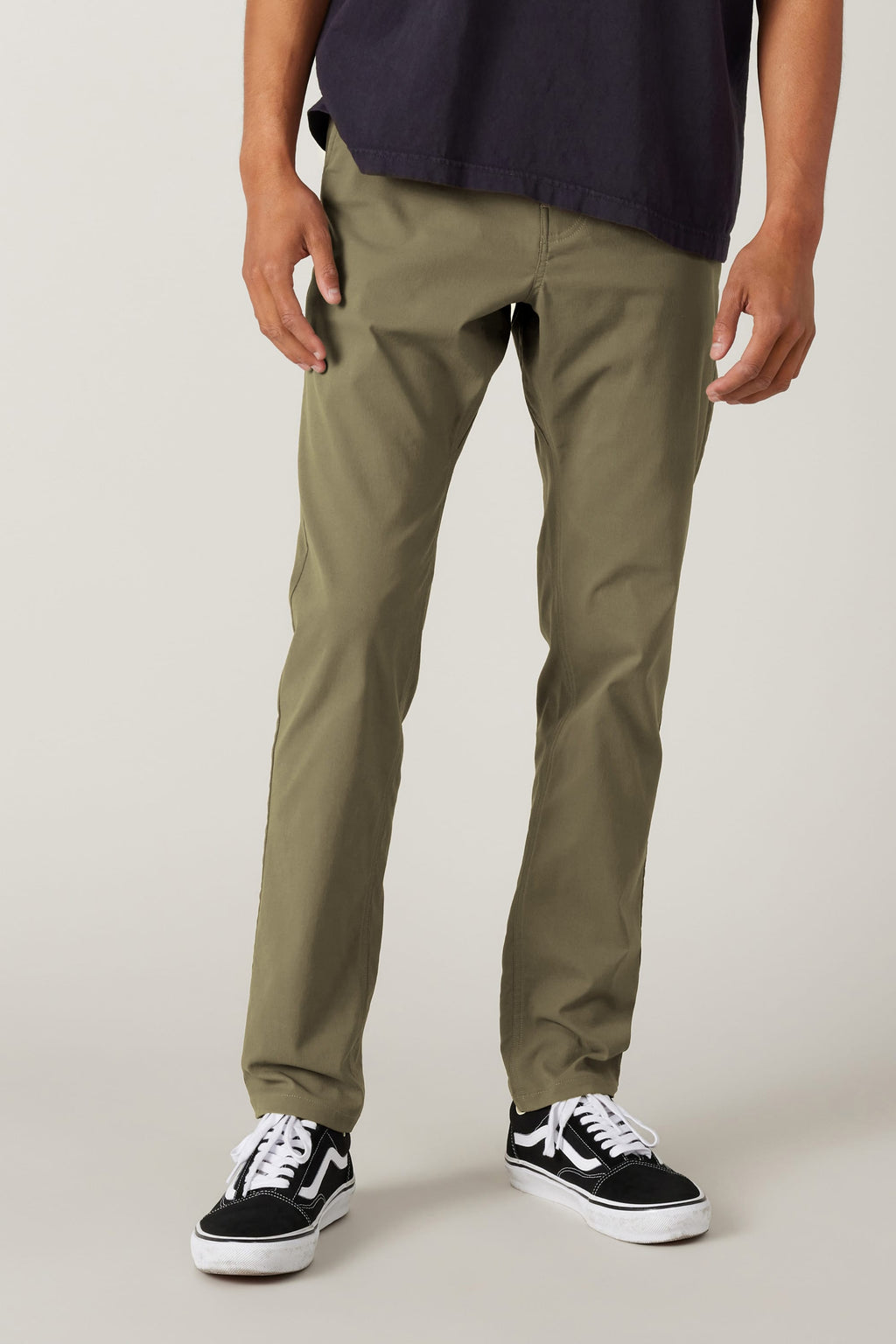 A person wearing 686 Men's Everywhere® Pant - Slim Fit in olive stands against a plain, light background. They pair the pants with a dark shirt and black-and-white sneakers; their hands are by their sides, and their face is not visible.