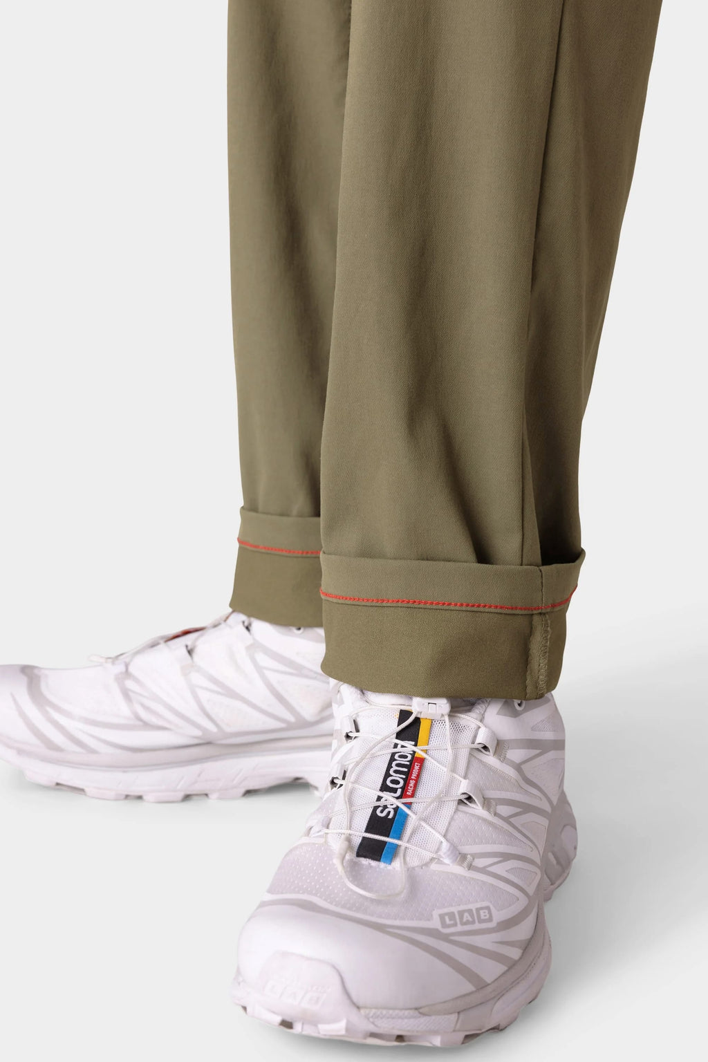 A close-up of a person wearing 686 Men's Everywhere® Pant - Slim Fit in olive green with red-stitched cuffs, paired with white Salomon sneakers featuring colorful branding on the tongue.