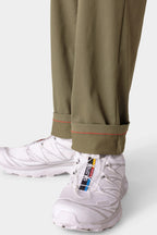 A close-up of a person wearing 686 Men's Everywhere® Pant - Slim Fit in olive green with red-stitched cuffs, paired with white Salomon sneakers featuring colorful branding on the tongue.