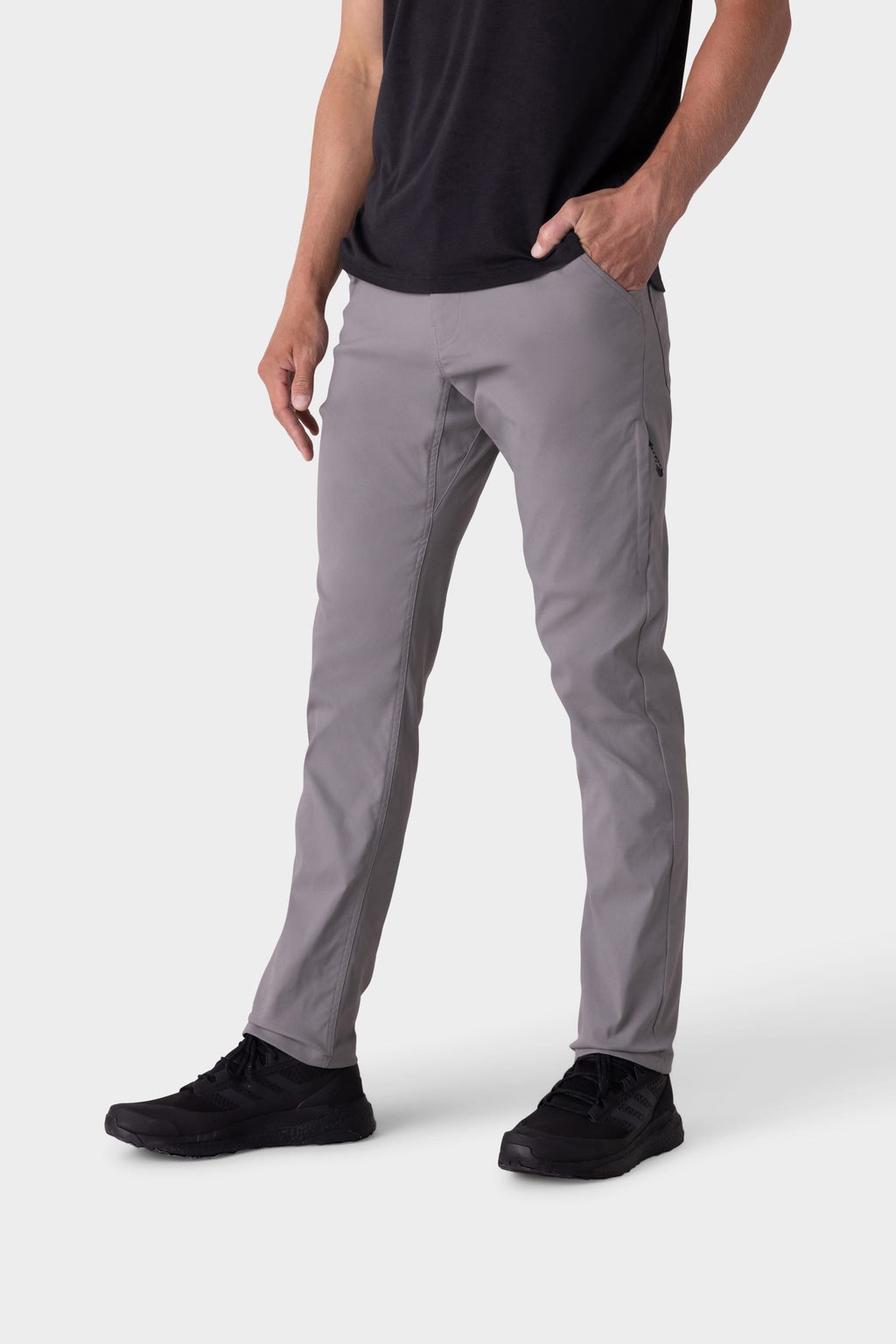 A person wearing 686 Men's Everywhere® Pant - Slim Fit in light gray, a black t-shirt, and black shoes stands with one hand in their pocket against a plain white background.