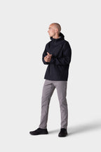 A person wearing a black hooded jacket, 686 Men's Everywhere® Pant - Slim Fit in light gray, and black shoes stands against a plain light gray background, looking to the side with hands clasped.
