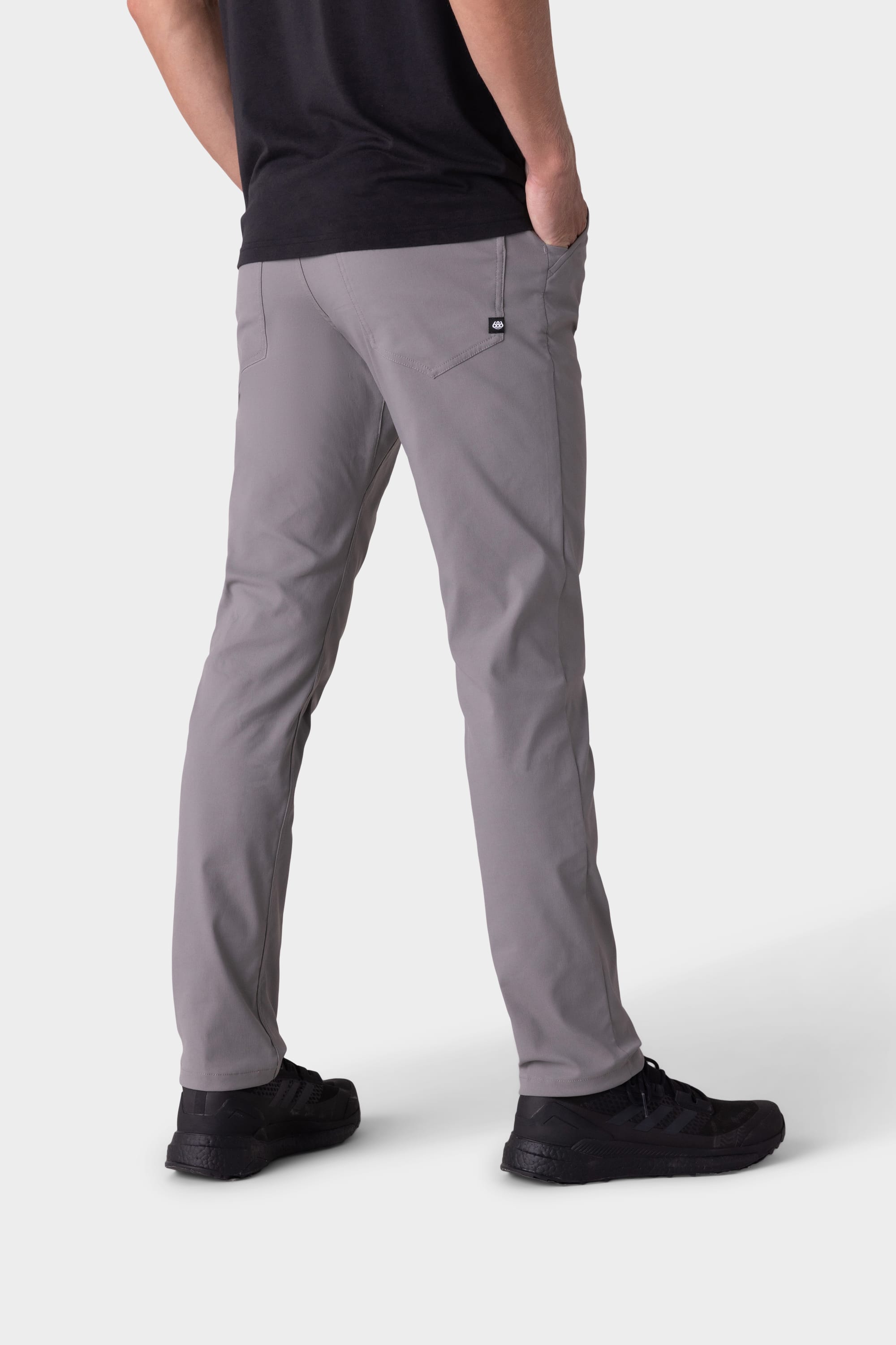 A person wearing 686 Men's Everywhere® Pant - Slim Fit in light gray, paired with a black shirt and black shoes, stands with hands in pockets facing away from the camera against a plain white background.