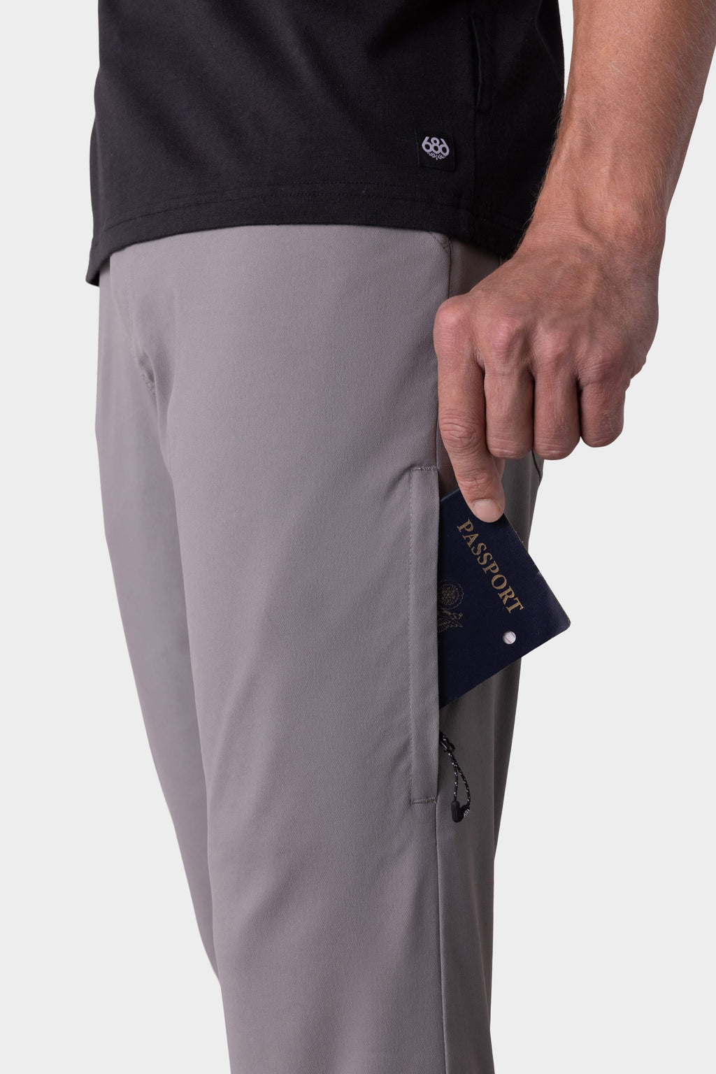 A person wearing stain-resistant, light gray 686 Men's Everywhere® Pant - Slim Fit and a black shirt slides a navy blue passport into the right front pocket. The image is cropped to show only their torso and hand.
