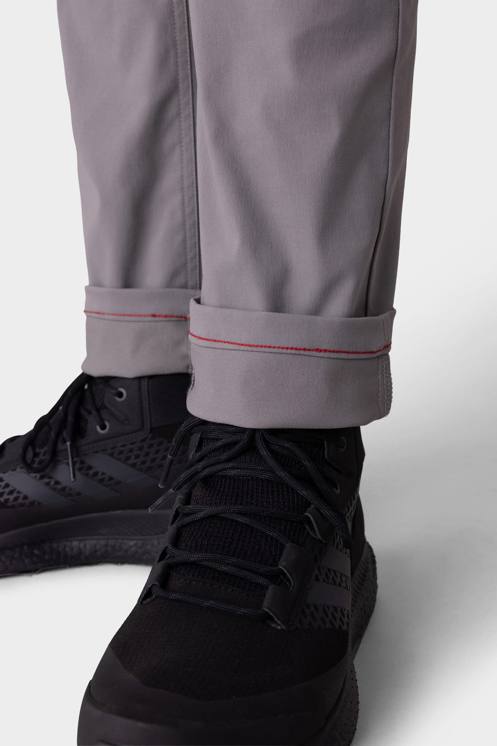 A close-up shows someone wearing 686 Men's Everywhere® Pant - Slim Fit in gray, cuffs rolled to display a red stitch detail. The breathable 686 pants are paired with black athletic sneakers.