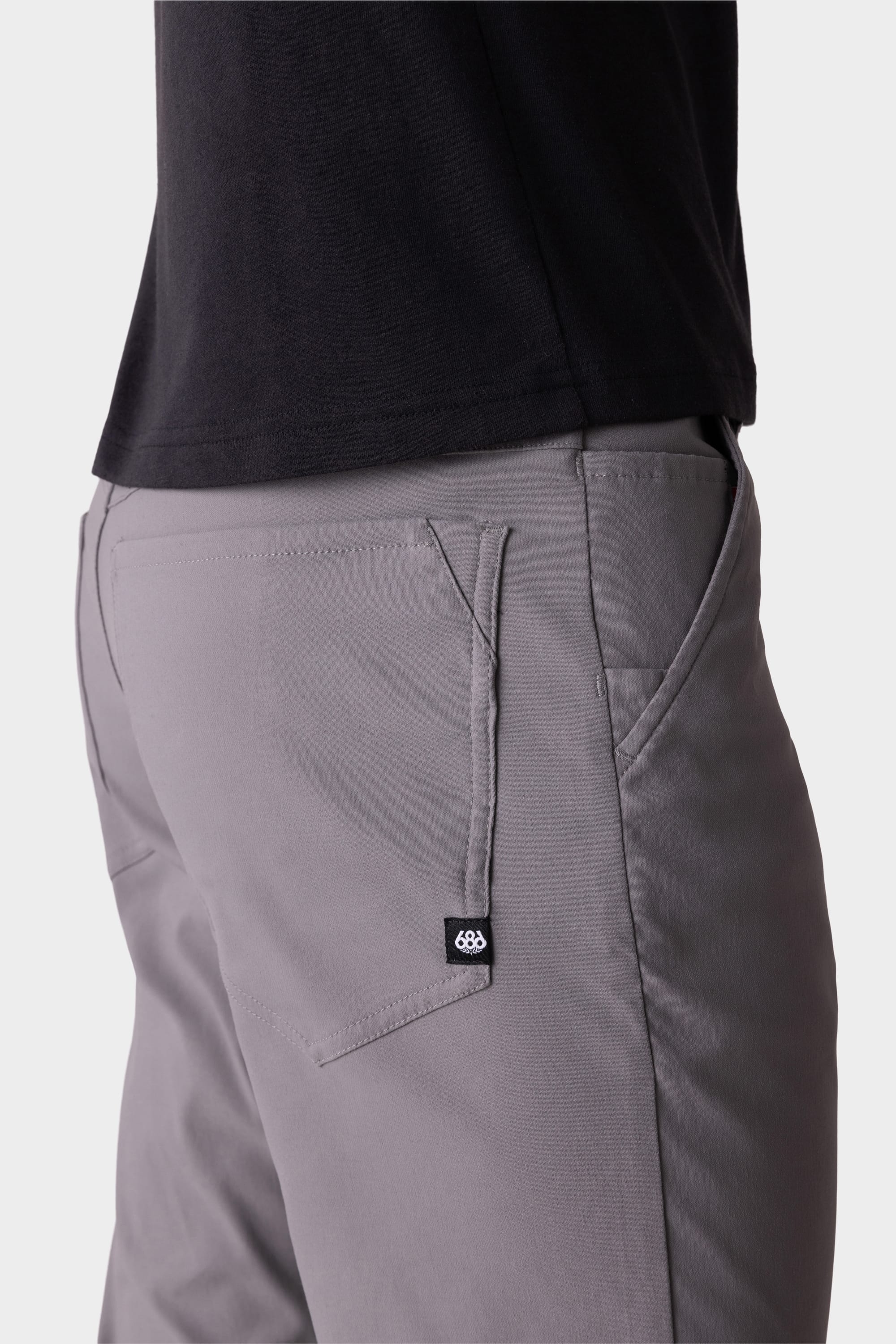 A close-up side view shows a person wearing 686 Men's Everywhere® Pant - Slim Fit in gray with a black shirt. The breathable 686 pants feature a small black tag with white logo near the back pocket, set against a plain light-colored background.