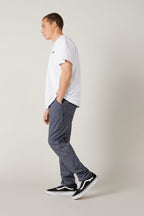 A man stands in profile against a plain background wearing a white T-shirt, 686 Men's Everywhere® Pant - Slim Fit in gray, and black Vans sneakers. His right hand is in his pocket and he looks slightly downward.