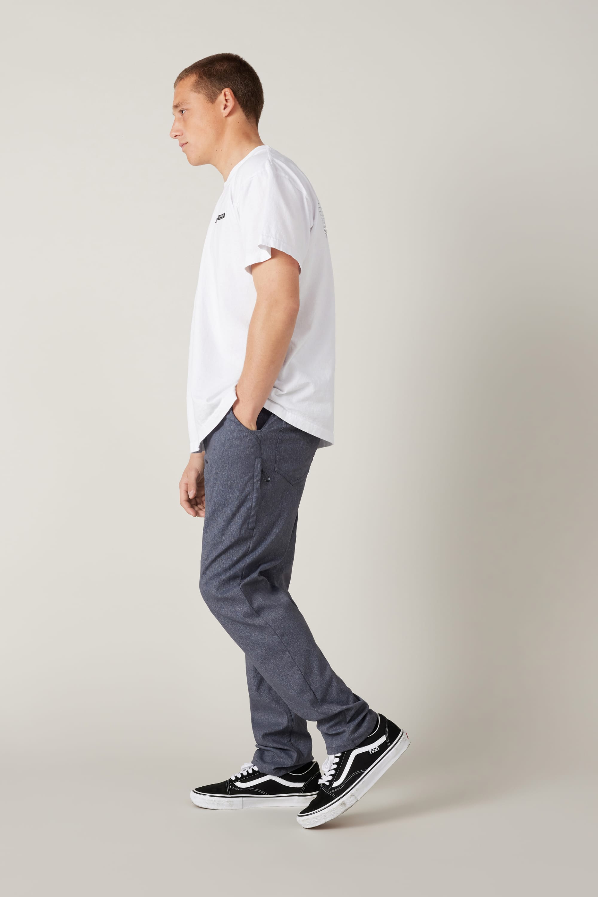 A man stands in profile against a plain background wearing a white T-shirt, 686 Men's Everywhere® Pant - Slim Fit in gray, and black Vans sneakers. His right hand is in his pocket and he looks slightly downward.