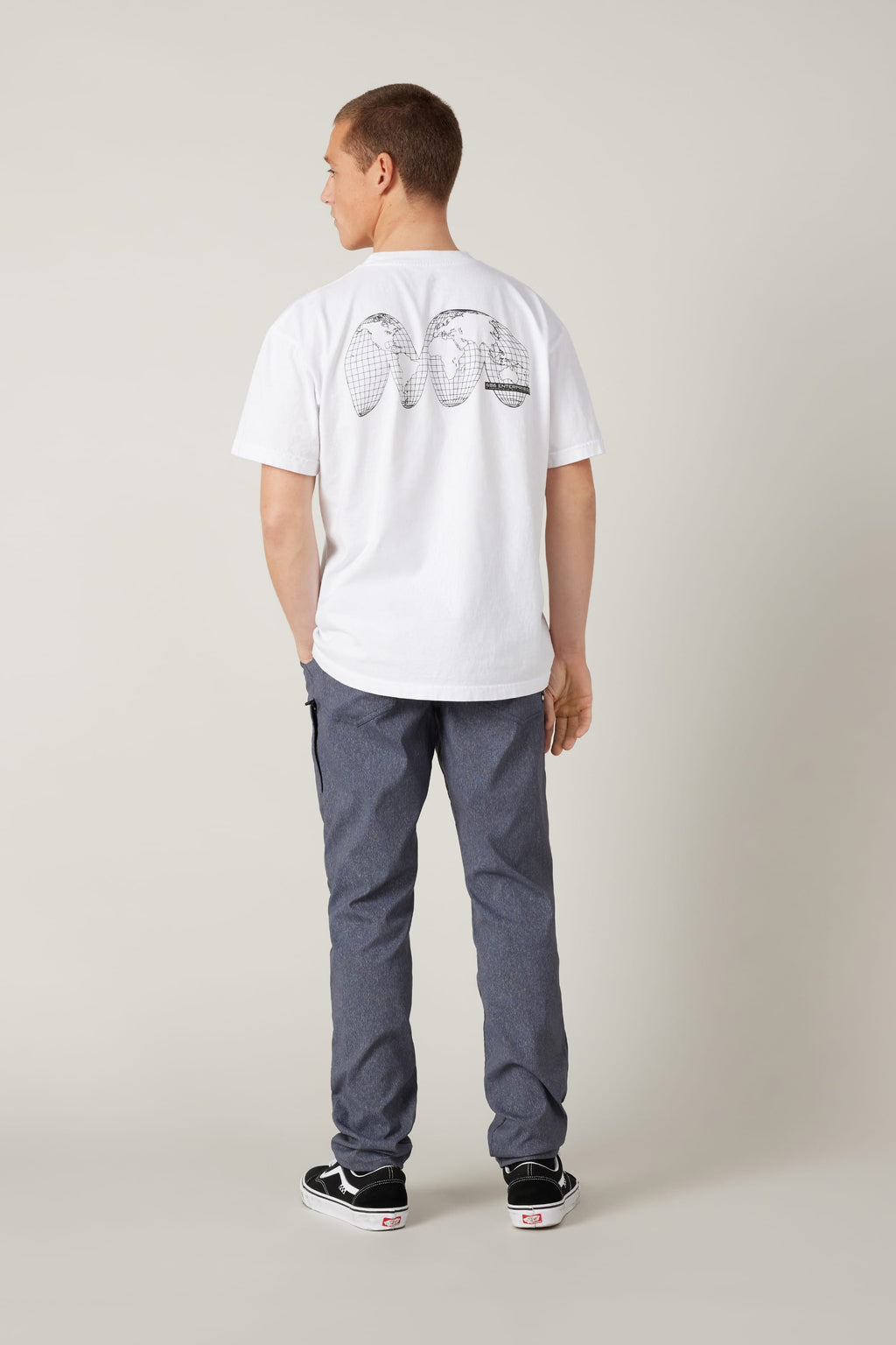 A person stands facing away, wearing a white T-shirt with black graphics, 686 Men's Everywhere® Pant - Slim Fit by 686, and black-and-white sneakers, against a plain light background.