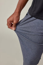 A person wearing 686 Men's Everywhere® Pant - Slim Fit in heathered blue and a black shirt stretches the fabric with their left hand, showcasing the pant’s impressive flexibility and stain-resistant material.