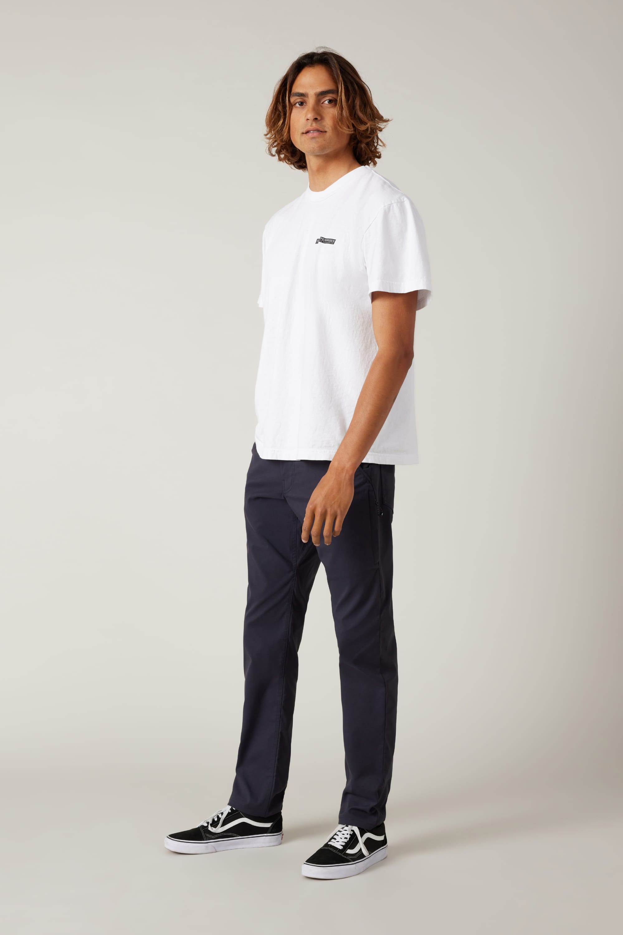 A young man with shoulder-length hair stands slightly to the side, wearing a white T-shirt, 686 Men's Everywhere® Pant - Slim Fit, and black sneakers with white soles against a plain light background.