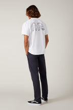 A person with wavy hair faces away, wearing a white tee with black graphics, 686 Men's Everywhere® Pant - Slim Fit in dark color, and black sneakers, all set against a plain light background.