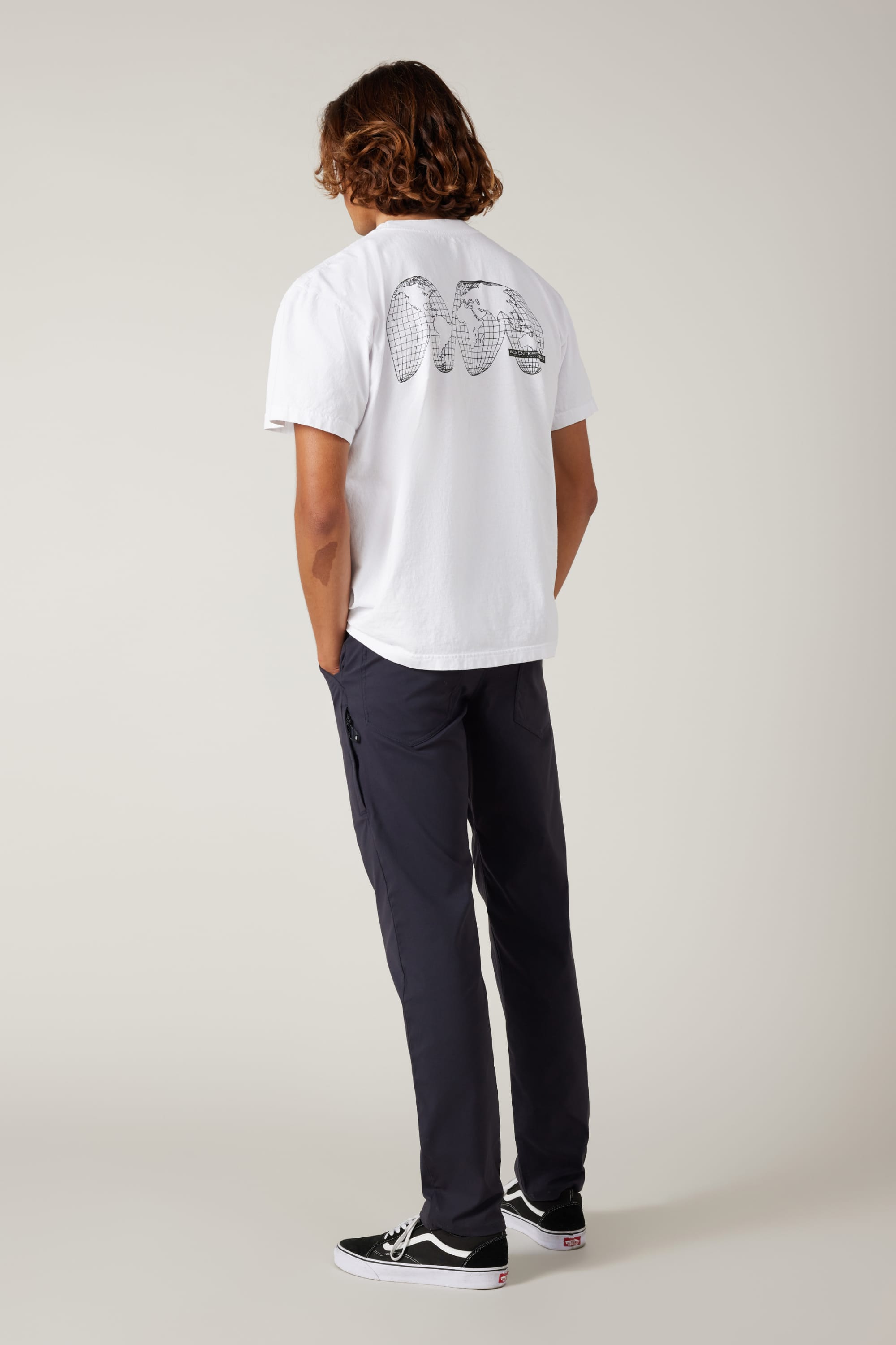 A person with wavy hair faces away, wearing a white tee with black graphics, 686 Men's Everywhere® Pant - Slim Fit in dark color, and black sneakers, all set against a plain light background.