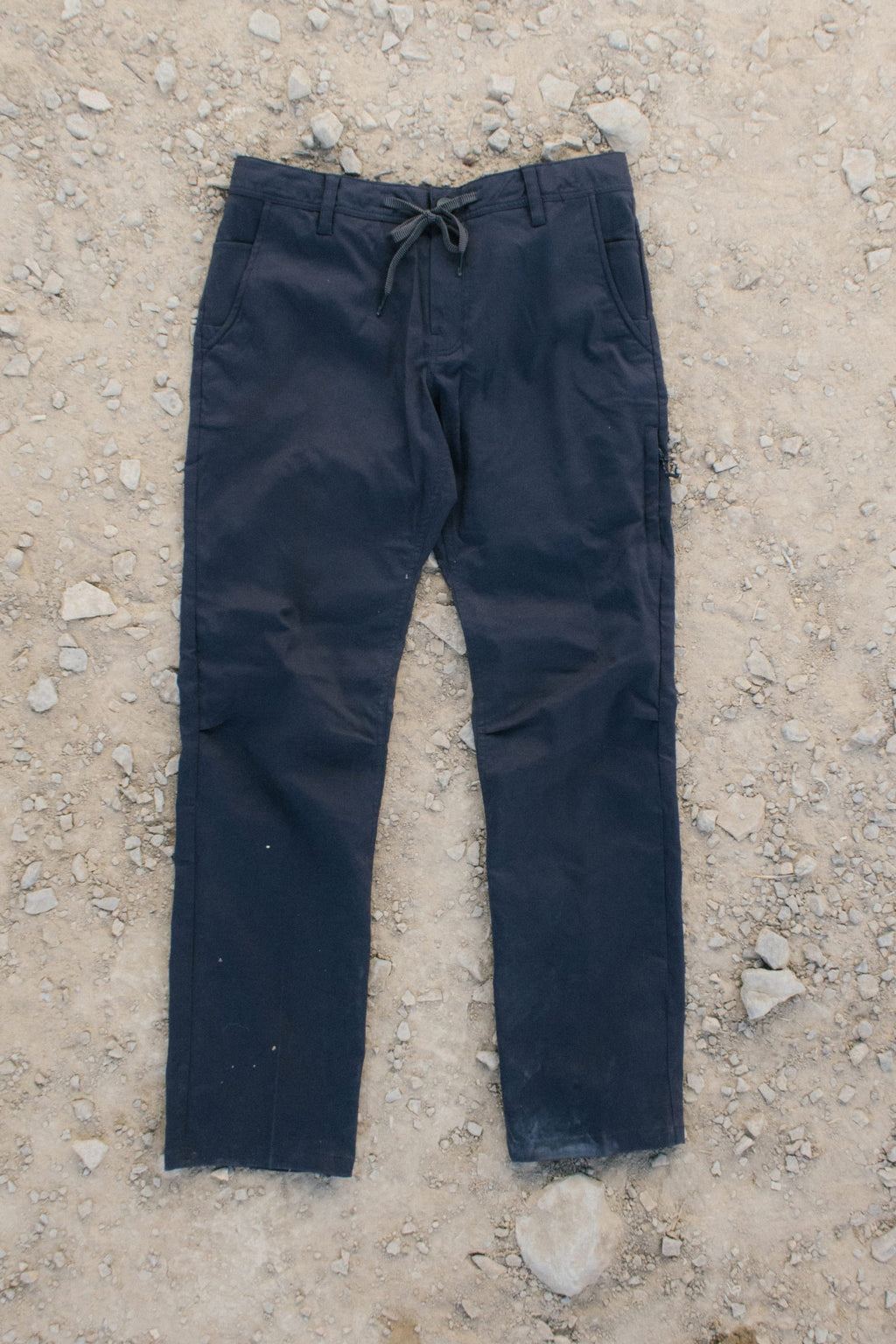 A pair of 686 Men's Everywhere® Pant - Slim Fit in black lies flat on rocky, sandy ground, shown with both legs extended and the waistband visible.