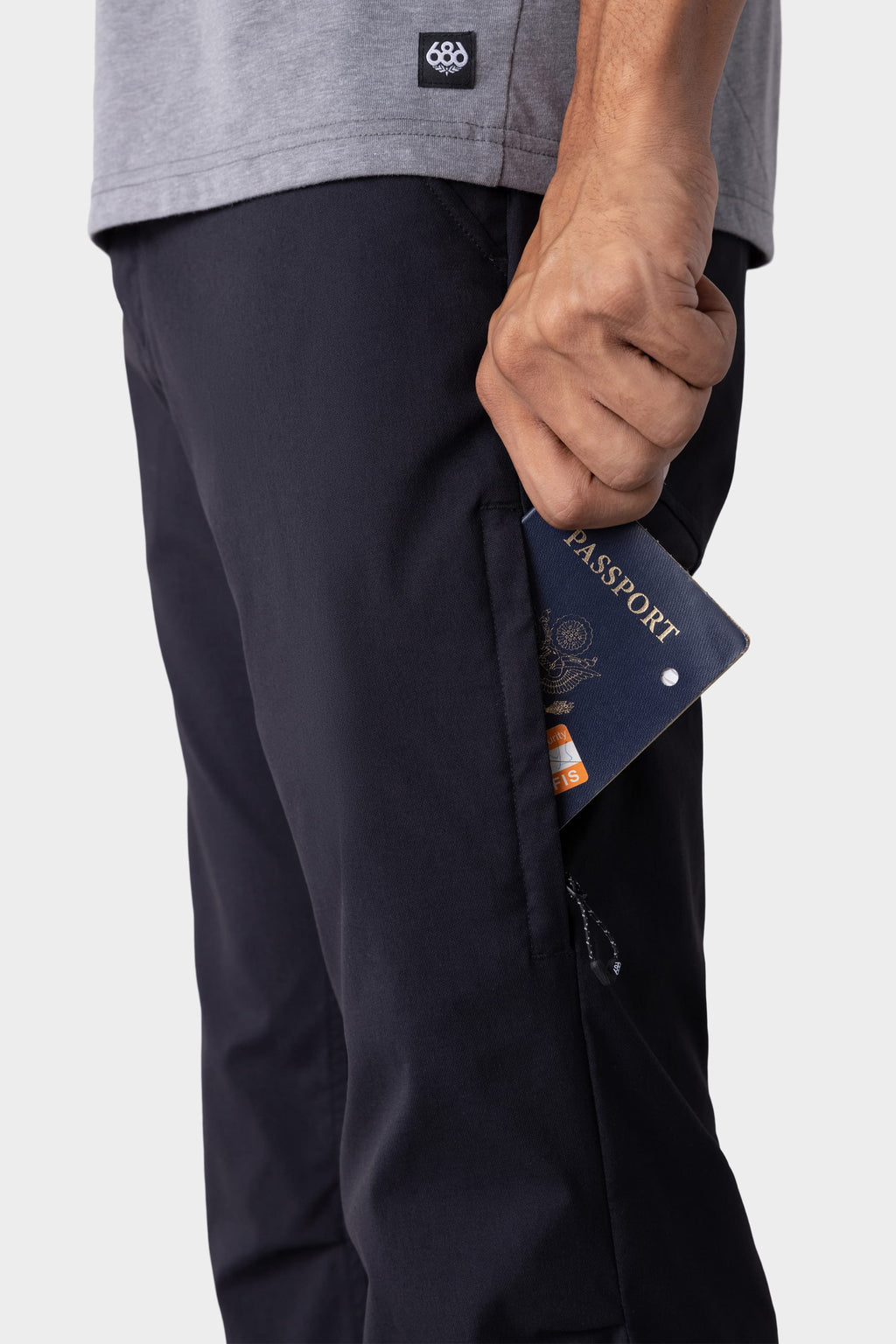 Wearing 686 Men's Everywhere® Pant - Slim Fit, a person in a gray shirt slides a passport and card into the side pocket. The cropped image highlights the hand, pocket, and passport.