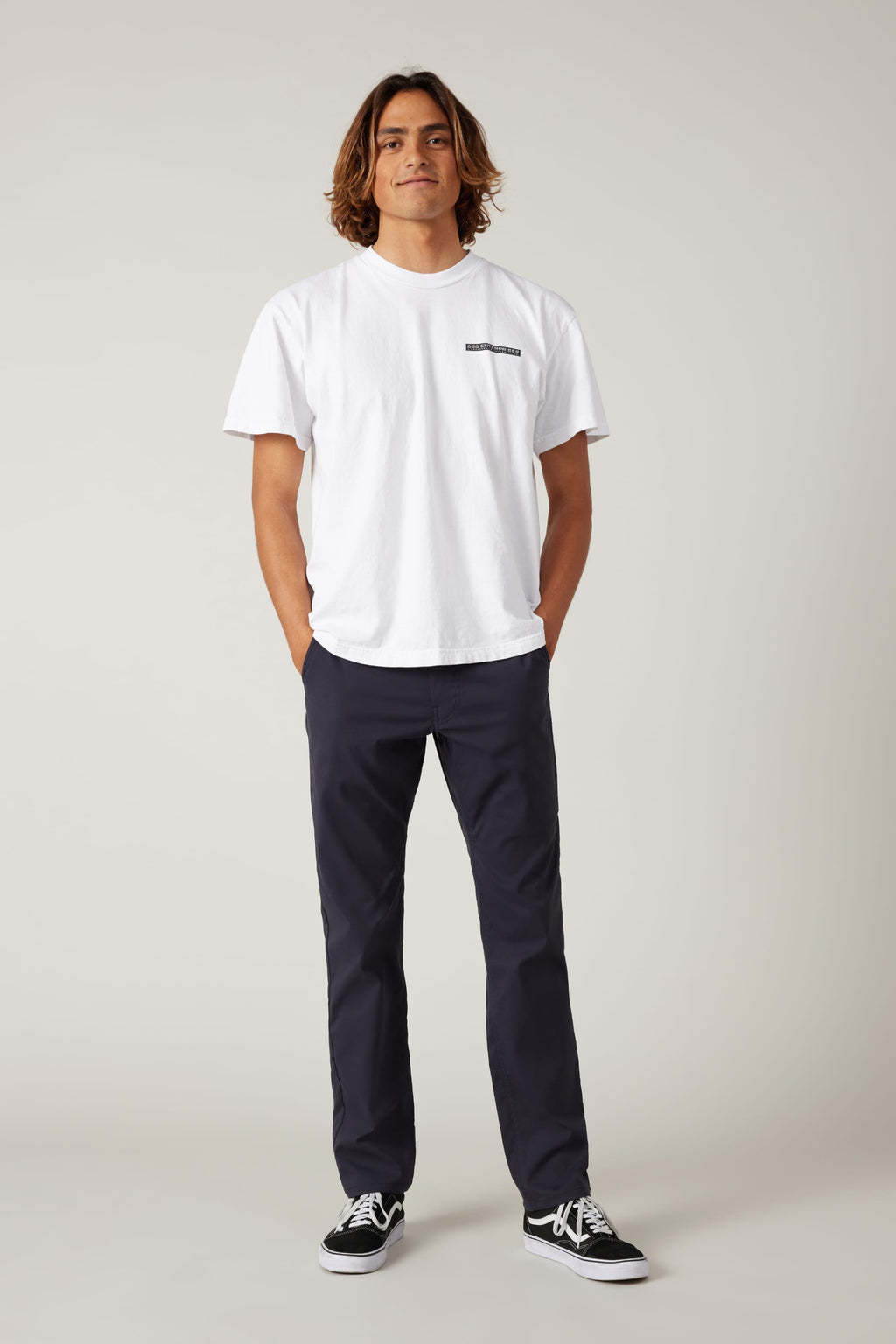 A person with shoulder-length hair stands facing forward, wearing a white t-shirt, 686 Men's Everywhere® Pant - Slim Fit in navy, and black sneakers, with hands in their pockets against a plain light background.