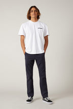 A person with shoulder-length hair stands facing forward, wearing a white t-shirt, 686 Men's Everywhere® Pant - Slim Fit in navy, and black sneakers, with hands in their pockets against a plain light background.