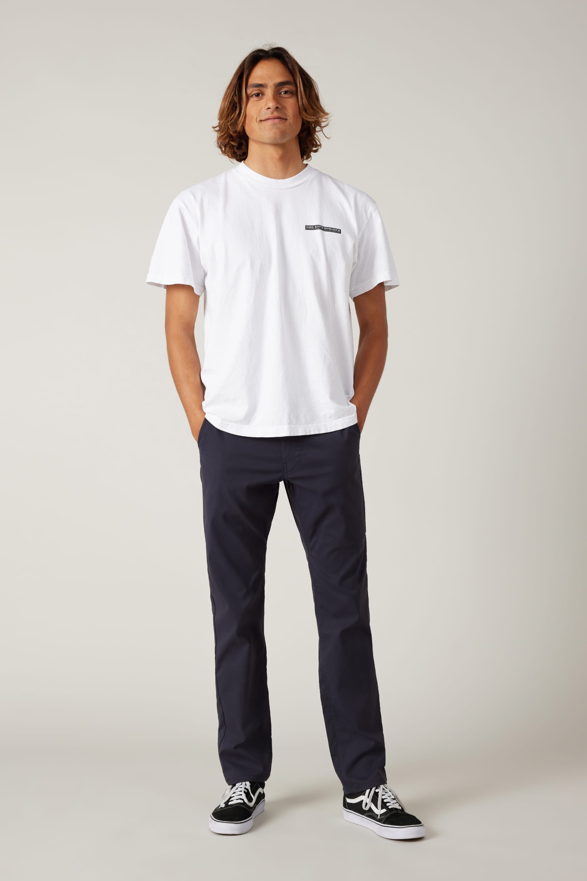 A person with shoulder-length hair stands facing forward, wearing a white t-shirt, 686 Men's Everywhere® Pant - Slim Fit in navy, and black sneakers, with hands in their pockets against a plain light background.