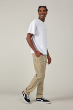 A man stands sideways, looking over his shoulder and smiling. He wears a white t-shirt, 686 Men's Everywhere® Pant - Slim Fit from 686 with subtle patterns, and black Vans sneakers against a plain light background.